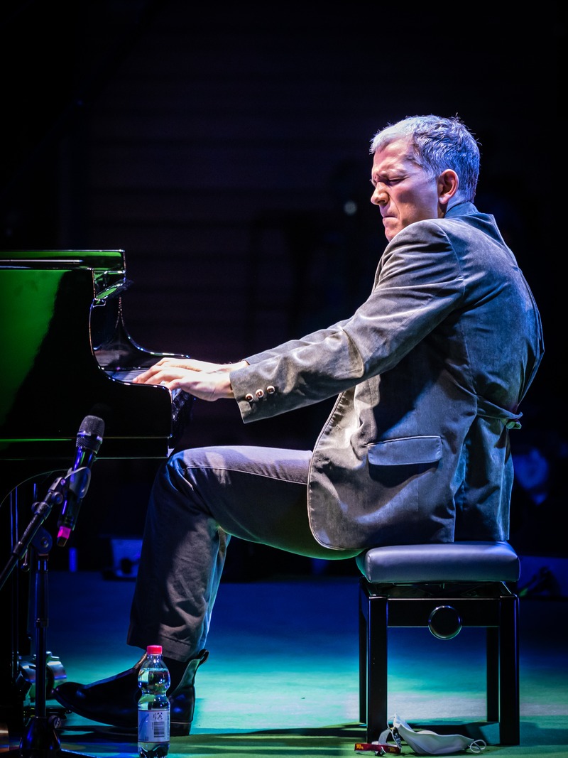 Brad Mehldau 身穿灰色西装,在三角钢琴前侧身弹奏