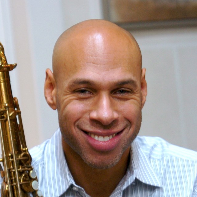 Joshua Redman 手持萨克斯的正面半身肖像，浅蓝条纹衬衫，微笑看向镜头